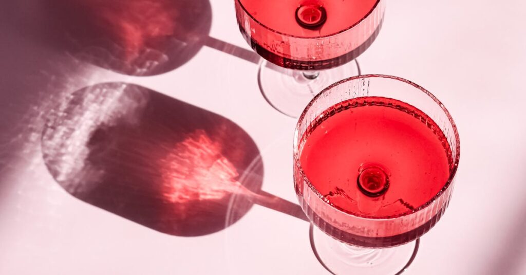 Two elegant glasses of red beverage casting artistic shadows on a pink surface in bright lighting.